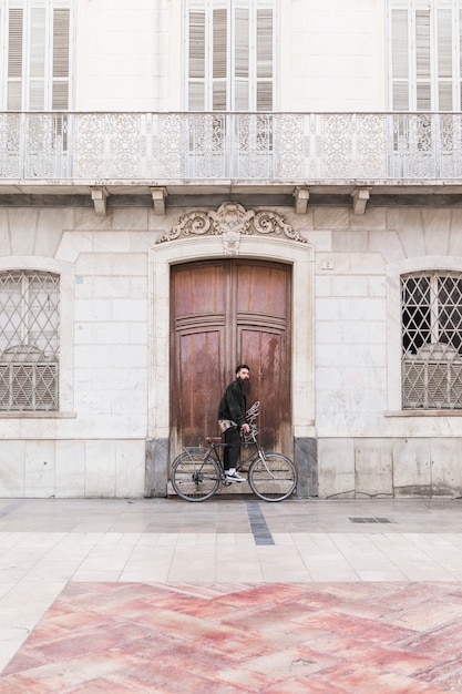 Бесплатное фото Молодой человек с велосипедом стоит перед старинным зданием