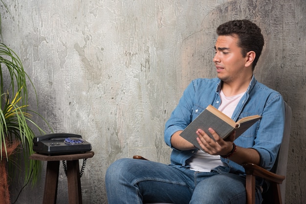 Молодой человек с книгой смотрит на телефон и сидит на стуле на мраморном фоне. Фото высокого качества