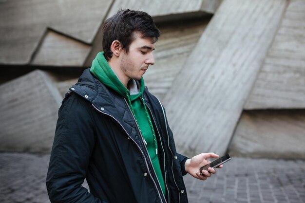 Young man with dark hair in black jacket standing outside with mobile phone in hands. Thoughtful boy in green sweatshirt standing on street and looking in his cellphone