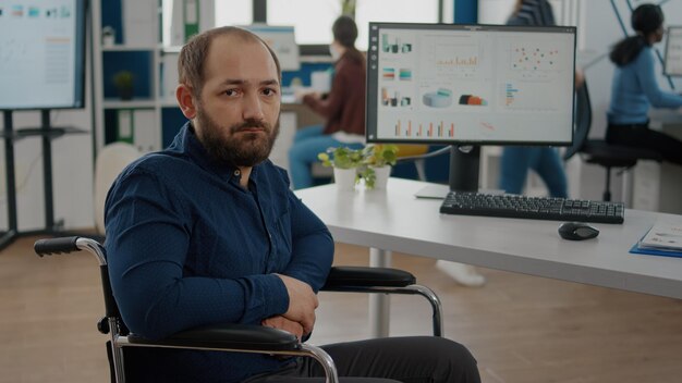 Young man with disabilities, invalid, disabled paralized handicapped immobilized looking at camera upset, tired sitting in wheelchair in business office room, working at financial project with team.