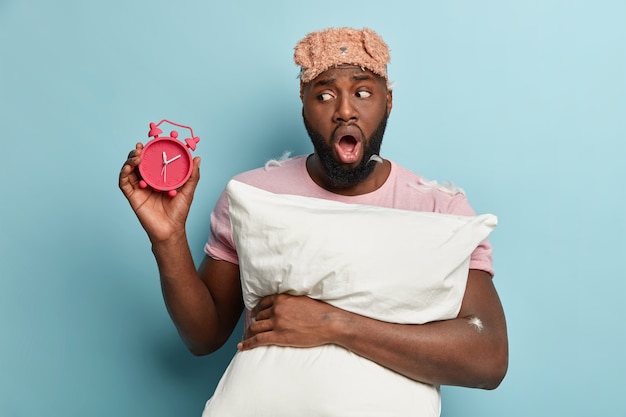 Young man with feathers on T-shirt holding pillow and alarm clock