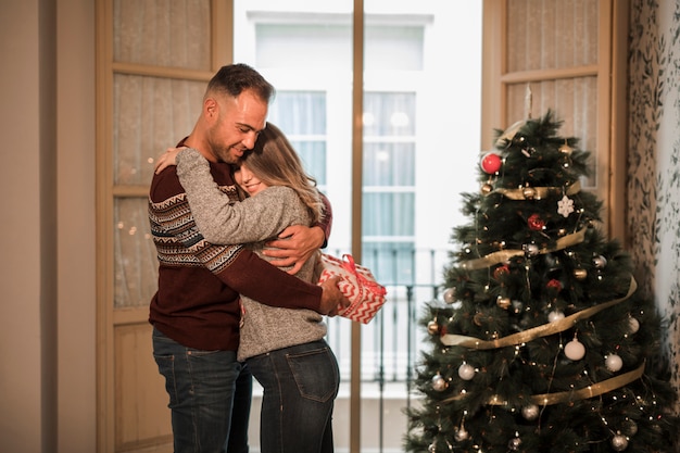 Giovane con scatola regalo abbracciando donna allegra vicino albero di natale