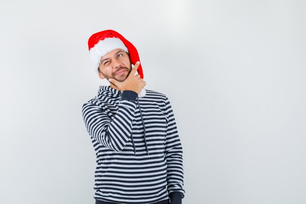 Молодой человек с рукой на подбородке, глядя вверх в толстовке с капюшоном, шляпе Санта-Клауса и глядя задумчиво, вид спереди.