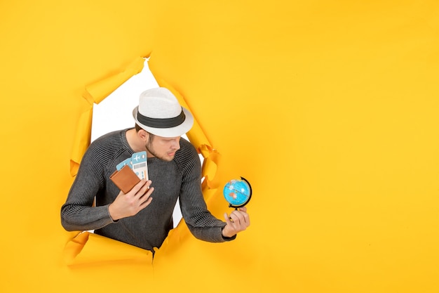 Young man with a hat holding foreign passport with ticket and small globe in a torn on yellow wall