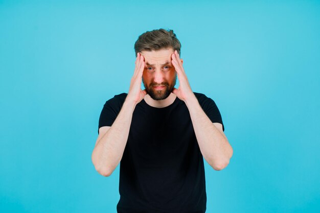 Young man with headache is putting hands on temples on blue background