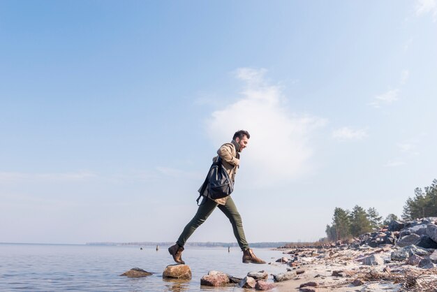 Young man with his backpack on shoulder jumping over the stones near the lake
