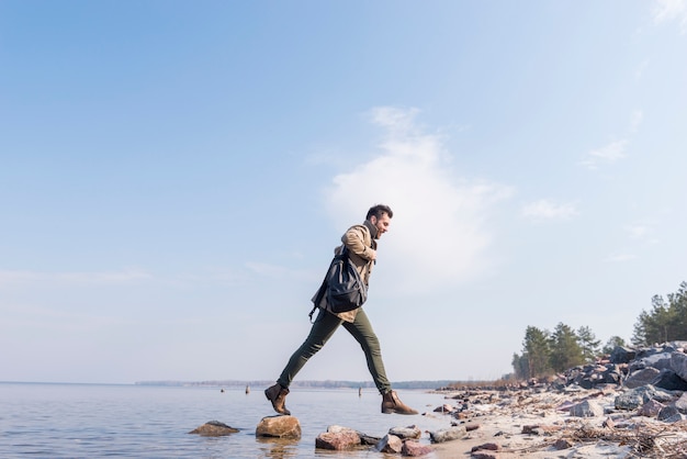 Young man with his backpack on shoulder jumping over the stones near the lake