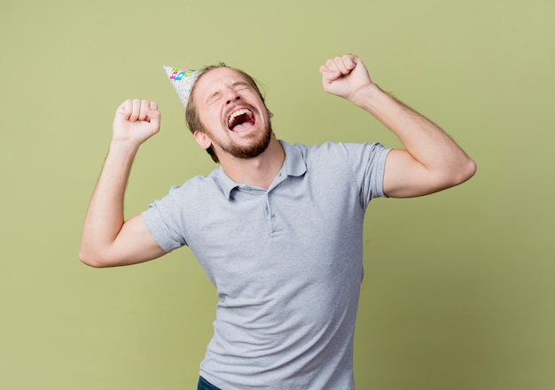 Молодой человек в праздничной кепке празднует день рождения, сумасшедший, счастливый и взволнованный над светлой стеной