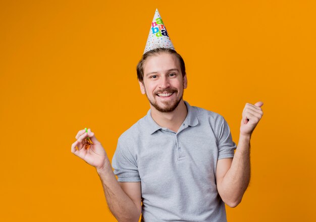 Молодой человек в праздничной кепке празднует день рождения, счастливый и взволнованный, улыбаясь, стоя над оранжевой стеной