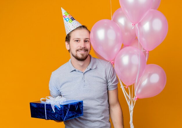 オレンジ色の上に幸せで陽気な風船と誕生日プレゼントの束を保持している誕生日パーティーを祝うホリデーキャップを持つ若い男