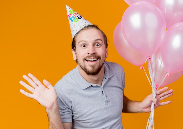 Young man with holiday cap celebrating birthday party holding bunch of balloons happy and excited smiling broadly  over orange