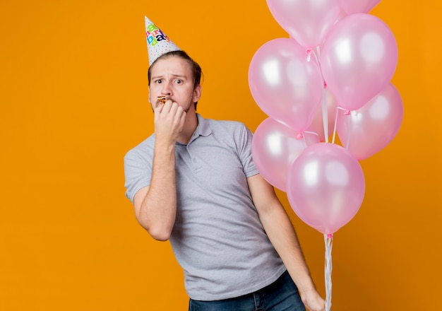Молодой человек в праздничной кепке празднует день рождения, держа кучу воздушных шаров над оранжевым