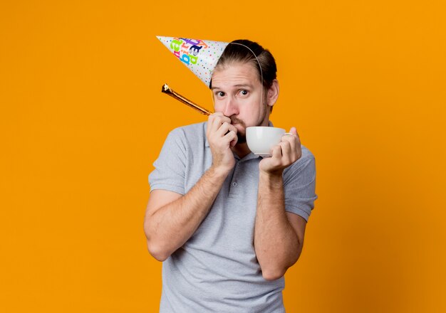 Young man with holiday cap celebrating birthday party holding coffee cap looking surprised and confused standing over orange wall