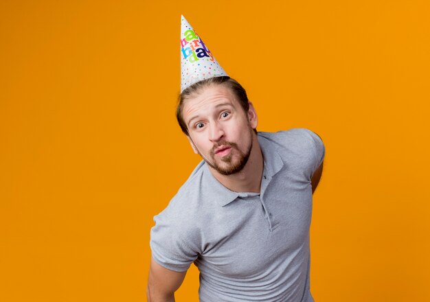 Young man with holiday cap celebrating birthday party keeping lips to kiss happy and cheerful standing over orange wall