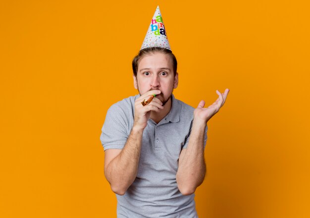Young man with holiday cap celebrating birthday party looking urprised standing over orange wall