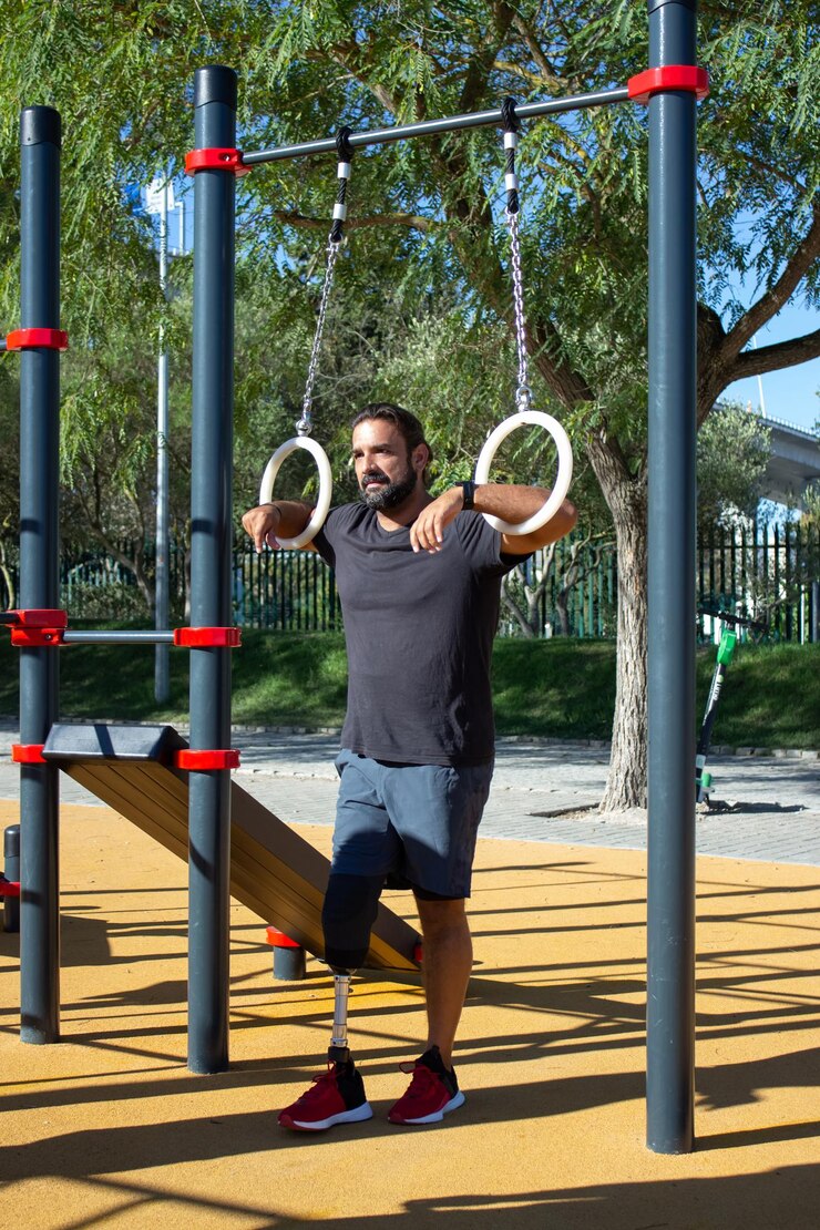 Free Photo young man with leg prothesis using gymnastic rings for training. strong sportsman standing on sports ground holding rings with hands looking far. health care, sports for people with disability concept