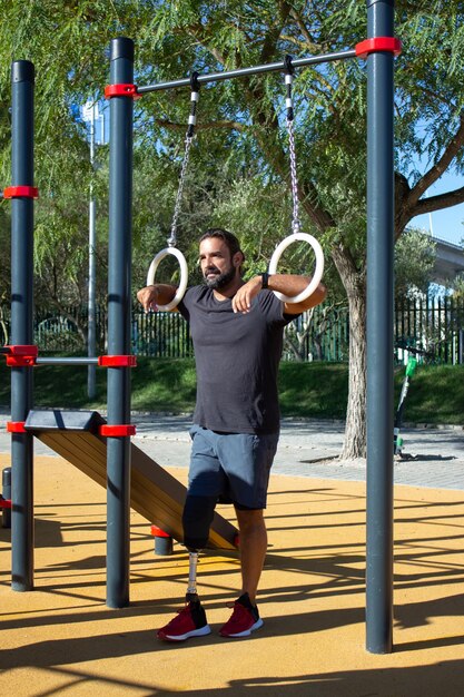 Young man with leg prothesis using gymnastic rings for training. Strong sportsman standing on sports ground holding rings with hands looking far. Health care, sports for people with disability concept