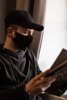 카페에서 까마귀 독서 잡지에 의료 보호 마스크와 검은 모자와 젊은 남자