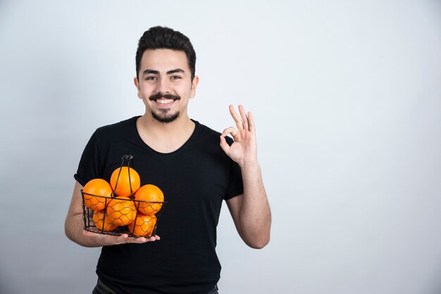 OKサインをしているオレンジ色の果物でいっぱいの金属製のバスケットを持つ若い男。