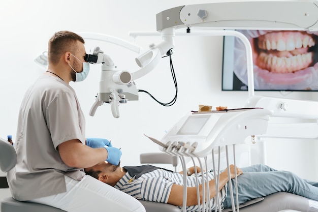 치과 의자에 턱받이가 있는 젊은 남자와 그 옆에 앉아 있는 치과의사. 그는 치과용 현미경을 사용하여 치아를 살펴보고 치과용 버와 거울을 들고 있습니다.