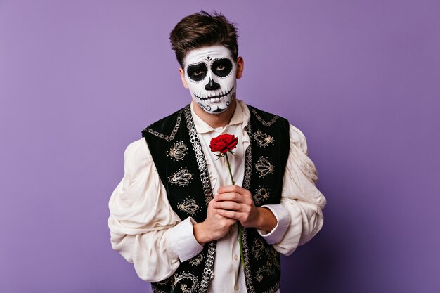 Молодой человек с грустным взглядом нежно держит на груди красный цветок. Портрет крупного плана брюнет в обмундировании Хэллоуина.