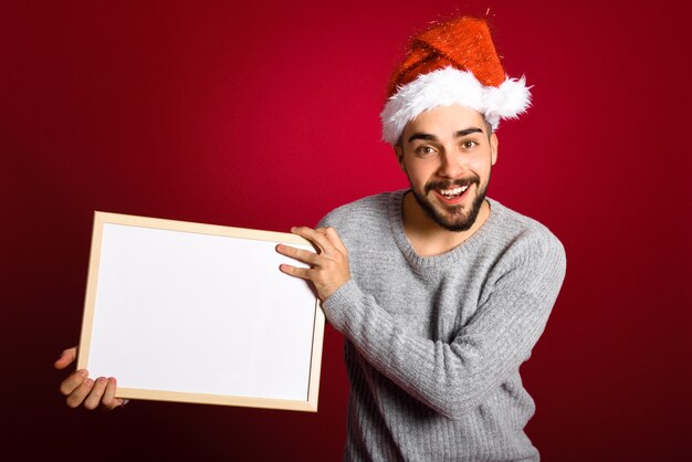 Молодой человек с Санта шляпу проведение пустой доски на красном фоне