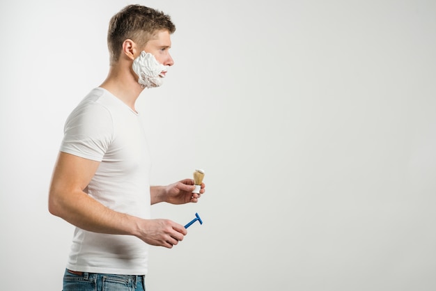 Молодой человек с пеной для бритья на щеках держит кисть и бритву на сером фоне