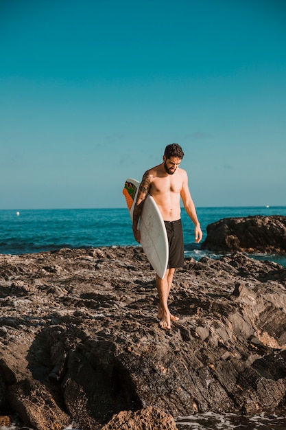 Young man with surf board going on stone shore near water