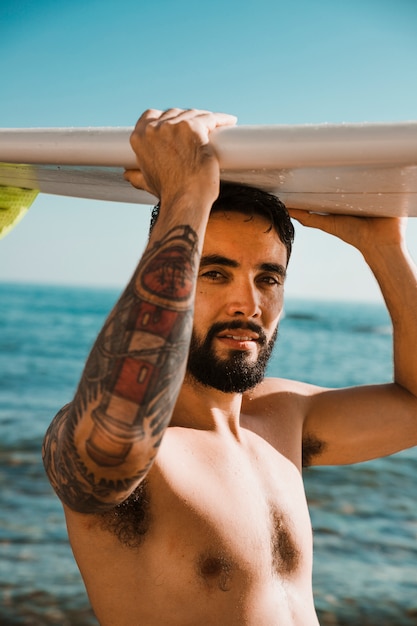 Young man with surf board on head on beach near water 