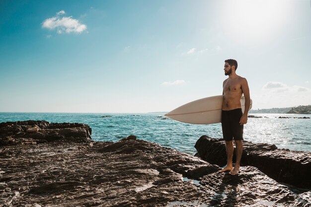 Young man with surf board on stone near sea