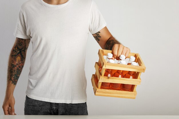 Young man with tattoos wearing jeans and a plain white t-shirt holds a wooden box with six unlabeled bottles of soft drinks