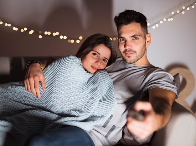 Young man with TV remote hugging woman and lying on sofa