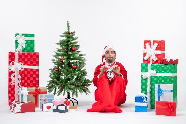 Young man with unsatisfied facial expreesion celebrate christmas holiday sitting in the ground and showing clock near gifts and decorated xmas tree