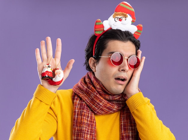 Бесплатное фото Молодой человек с теплым шарфом на шее в желтой водолазке и очках с забавной оправой на голове с рождественскими игрушками встревоженно стоит над фиолетовой стеной