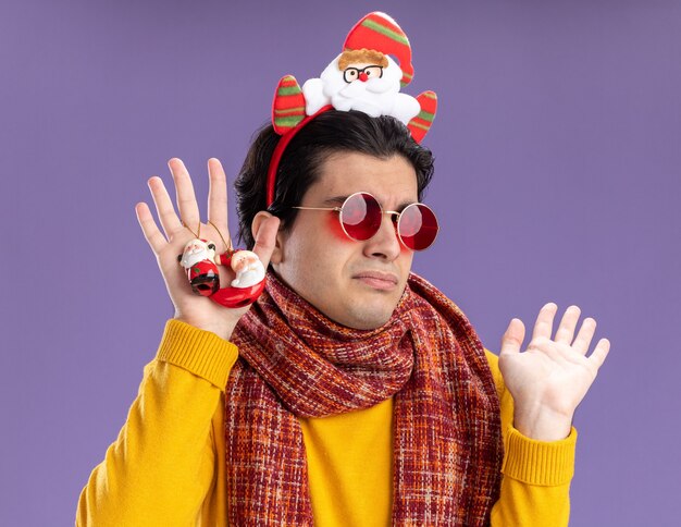 노란색 터틀넥 목에 따뜻한 스카프와 보라색 벽 위에 서있는 의심을 갖는 혼란스러워 보이는 크리스마스 장난감을 들고 머리에 재미있는 테두리가있는 안경을 가진 젊은 남자