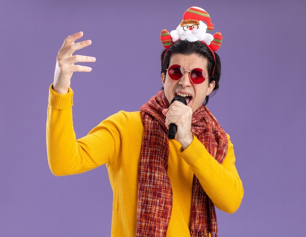 Young man with warm scarf around neck in yellow turtleneck and glasses with funny rim on head holding microphone happyand excited singing standing over purple wall