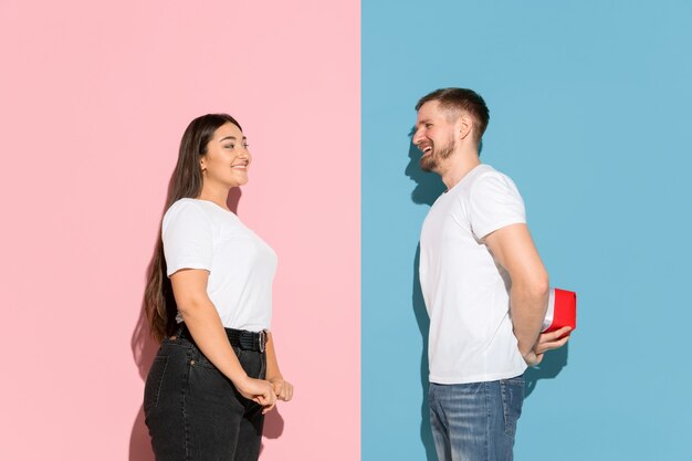 Young man and woman in casual clothes on pink, blue bicolored background. Man giving a gift to a woman