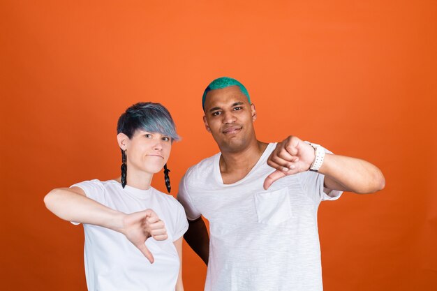 Young man and woman in casual white on orange wall both unhappy shows thumb down