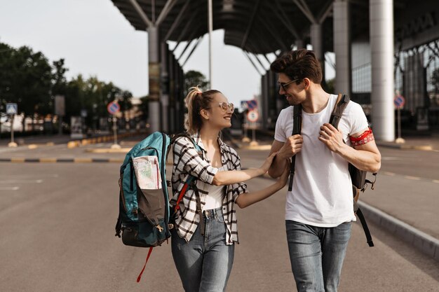 Young man and woman in jeans and white shirts talks and walks near airport Travelers with backpacks smiles and moves