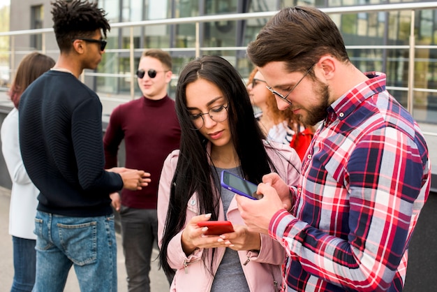 若い男と女の屋外の後ろに立っている彼らの友人とスマートフォンを使用して