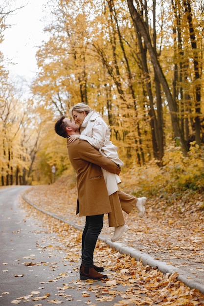 Young man and woman walking outside wearing beige coats. Blond woman and brunette man in autumn forest. Romantic couple kissing.