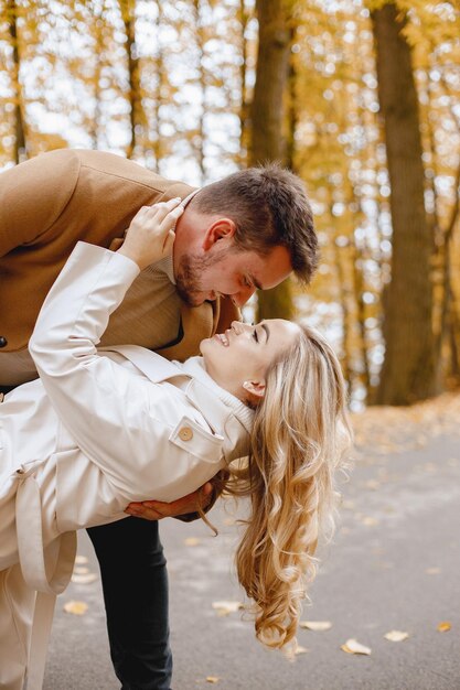 Young man and woman walking outside wearing beige coats. Blond woman and brunette man in autumn forest. Romantic couple kissing.