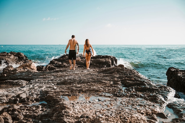 Young man and woman with surf boards going on stone shore to water