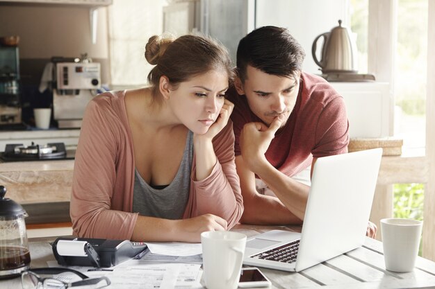 Young man and woman working together on laptop, paying utility bills via internet or using online mortgage calculator to save money on home loan, looking at screen with serious concentrated expression