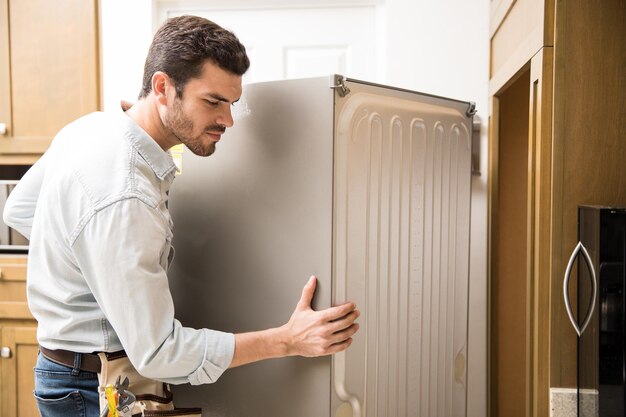 Молодой человек, работающий электриком, обнажает заднюю часть холодильника, чтобы проверить и отремонтировать его