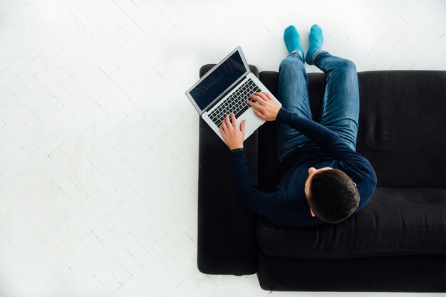 Young man working on laptop, sitting on black sofa, white floor. 