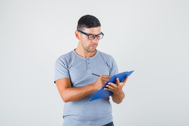 Young man writing on clipboard with pencil in grey t-shirt, glasses