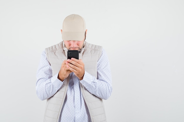 Молодой человек пишет тексты на мобильном телефоне в бежевой куртке и кепке и смотрит сосредоточенным. передний план.