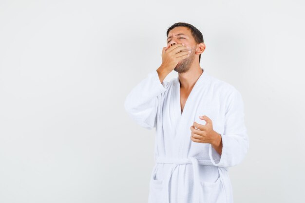 Young man yawning in white bathrobe and looking sleepy , front view.