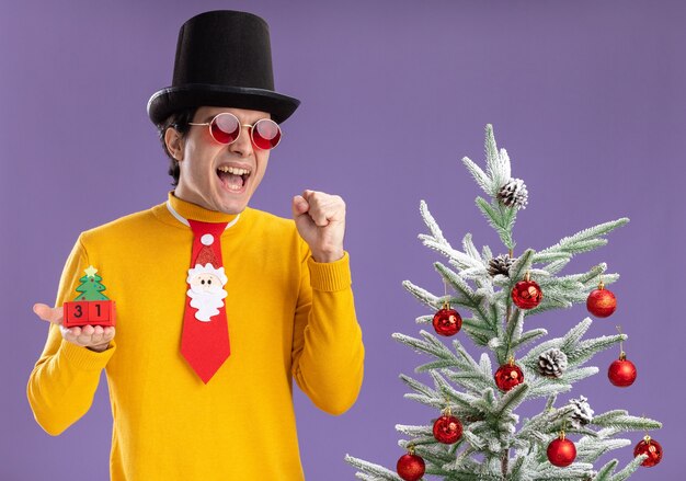 Young man in yellow turtleneck and glasses wearing black hat and funny tie holding cubes with new year date excited and happy standing next to a christmas tree over purple background